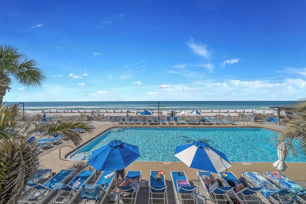 A pool area with lounge chairs and umbrellas, a clear sky, and a beach in the background.