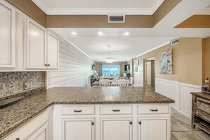 A kitchen with white cabinetry, granite countertops, and a view to the dining area. - Regency Towers 214 Heated Pool, Tiki Bar, Gym, FREE Beach Service, Golf&Perks (Panama City Beach)