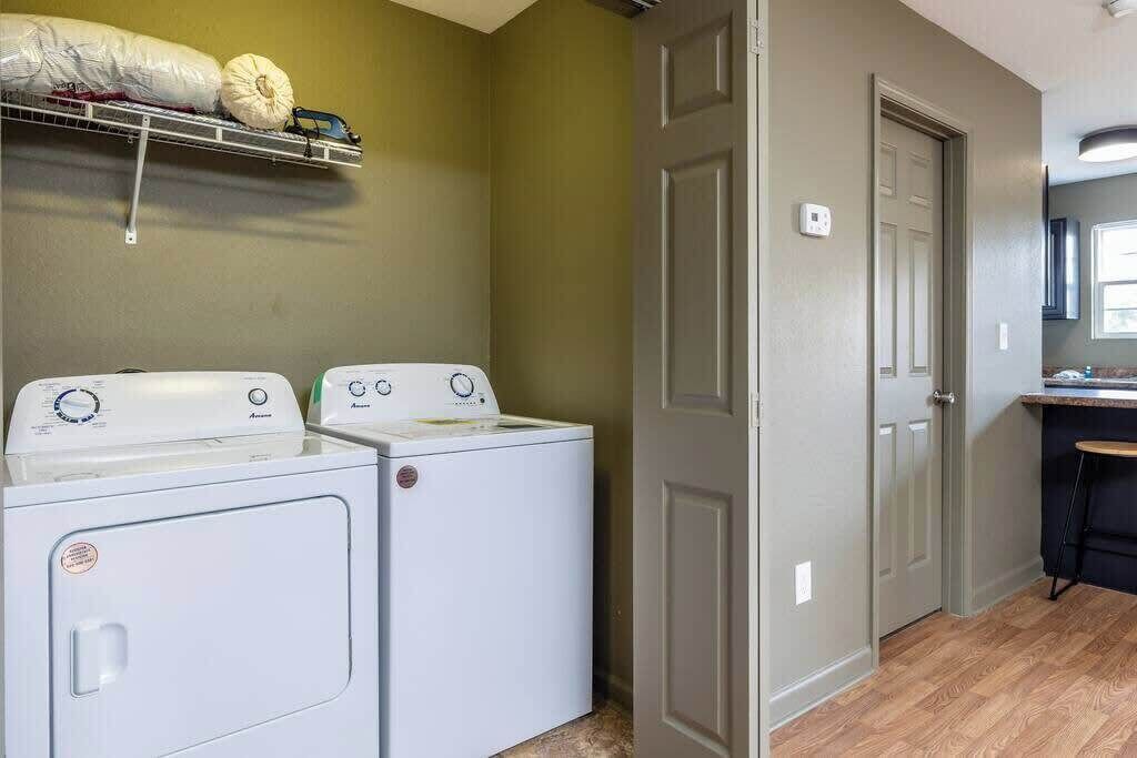 A laundry area with two white washing machines, a shelf with laundry items, and a door leading to a kitchen area.