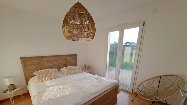 A bedroom with a wooden bed, a woven ceiling light, a wicker chair, and a sliding glass door leading to a patio. - Maison Bord de mer Avec Jardin Clos (Ploudalmézeau)