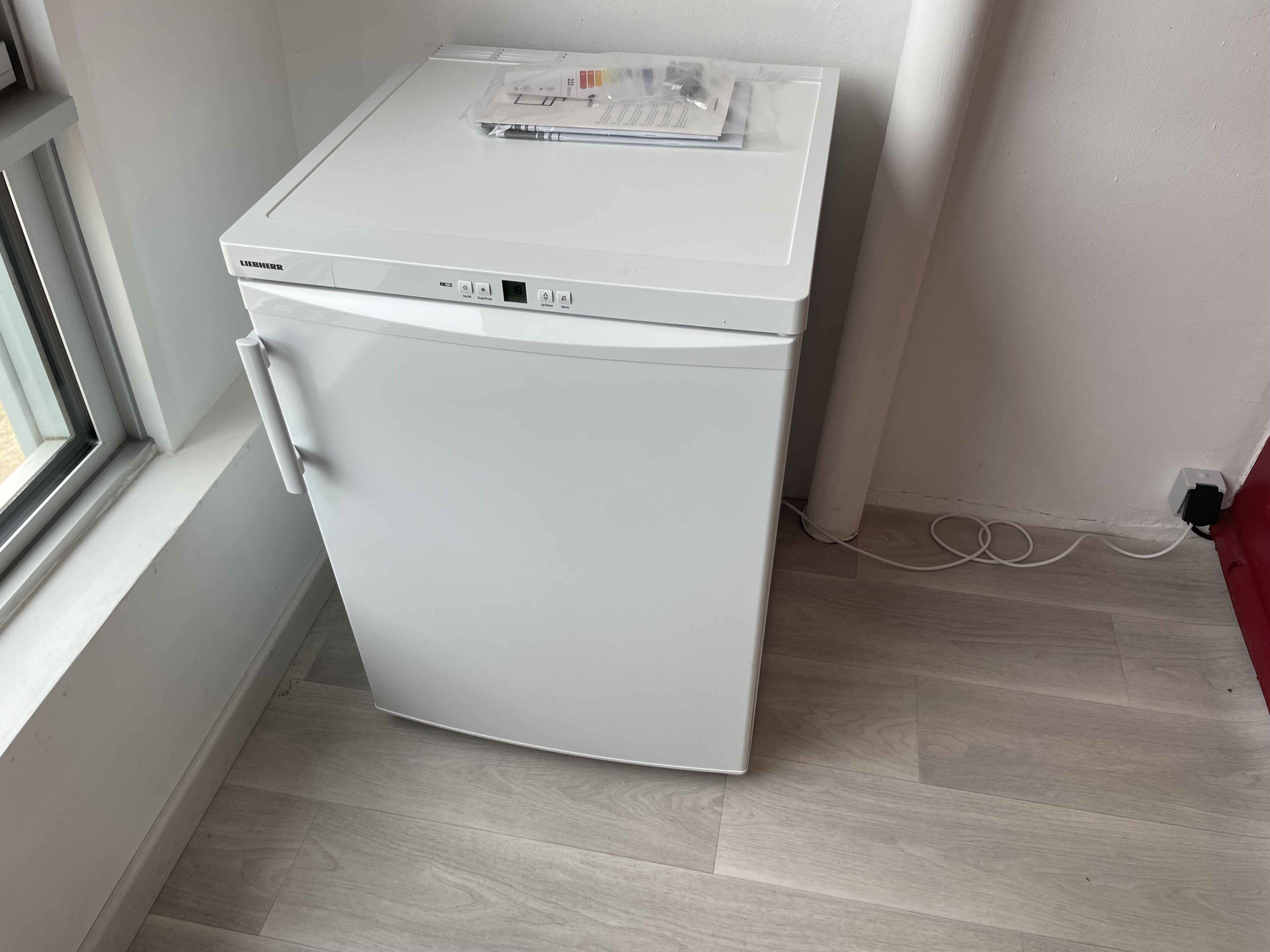 A compact white appliance, likely a refrigerator, in a corner with a window and a power outlet.