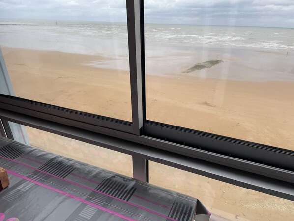 A view of a beach through a window, with a sandy shore and ocean waves visible. - Petit Cocon Courseullais with 2-star sea view (Courseulles-sur-Mer)