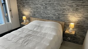 A bedroom with a white bedspread, a stone-patterned wall, and two bedside lamps. - Petit Cocon Courseullais with 2-star sea view (Courseulles-sur-Mer)