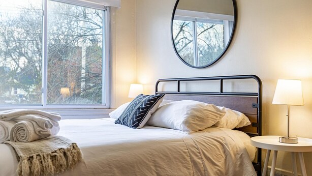 A neatly made bed with a white comforter and pillows, a round mirror above it, and a bedside table with a lamp. - Large 4BR Stadium Getaway, Walk to the Big House! (Ann Arbor)