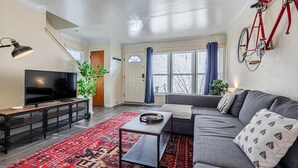 A modern living room with a large flat-screen TV, a gray sofa, a red patterned rug, and a bicycle mounted on the wall. - Large 4BR Stadium Getaway, Walk to the Big House! (Ann Arbor)