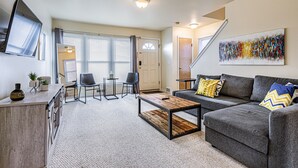 A modern living room with a grey sofa, a coffee table, and a TV mounted on the wall. There are two chairs and a small table with a plant. - Large 4BR Stadium Getaway, Walk to the Big House! (Ann Arbor)