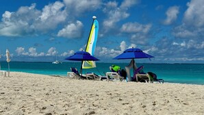 Una playa con cielo despejado, un velero a lo lejos y sillas de playa bajo sombrillas.