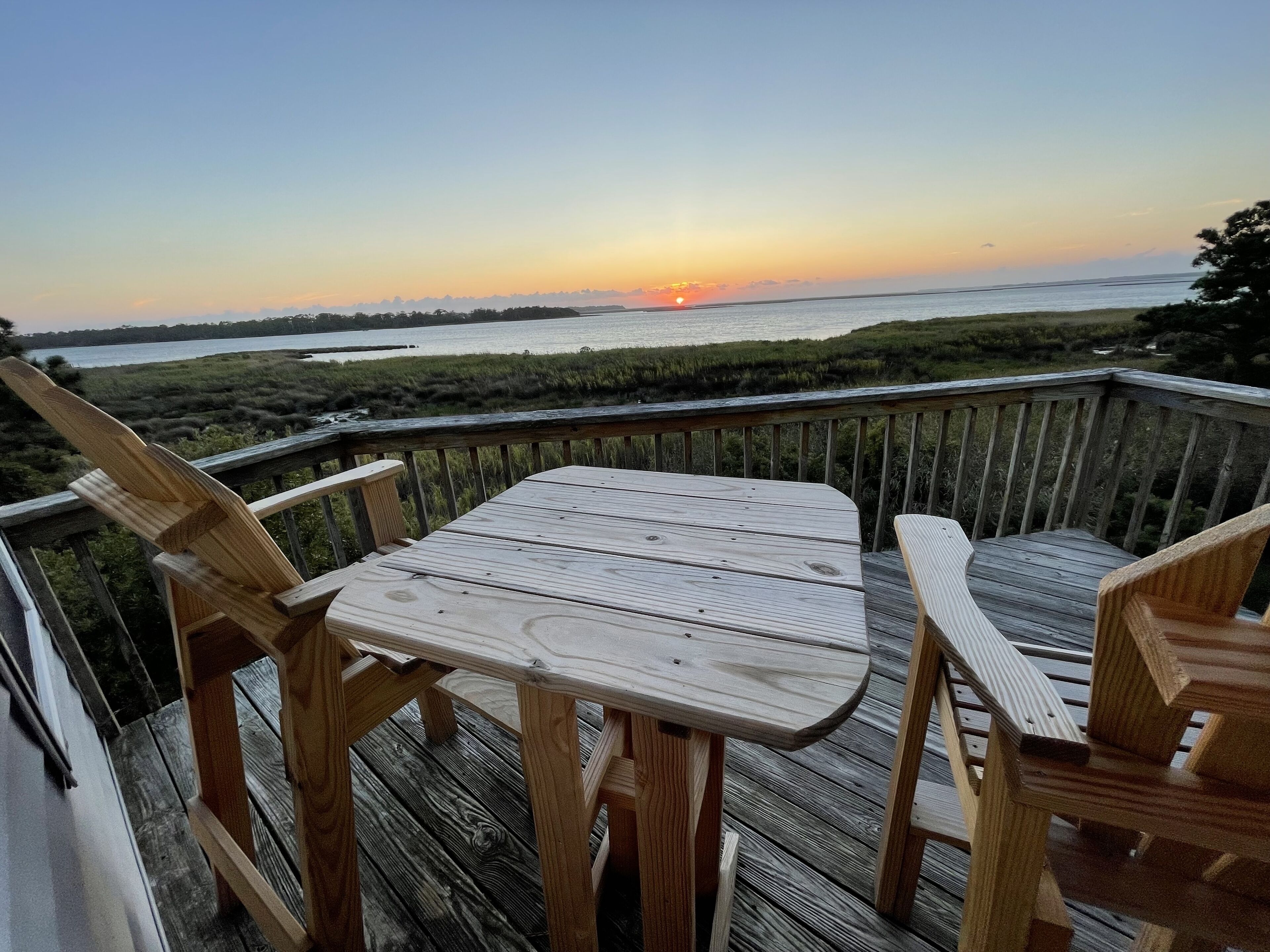A wooden deck with a table and chairs overlooking a sunset over a body of water.