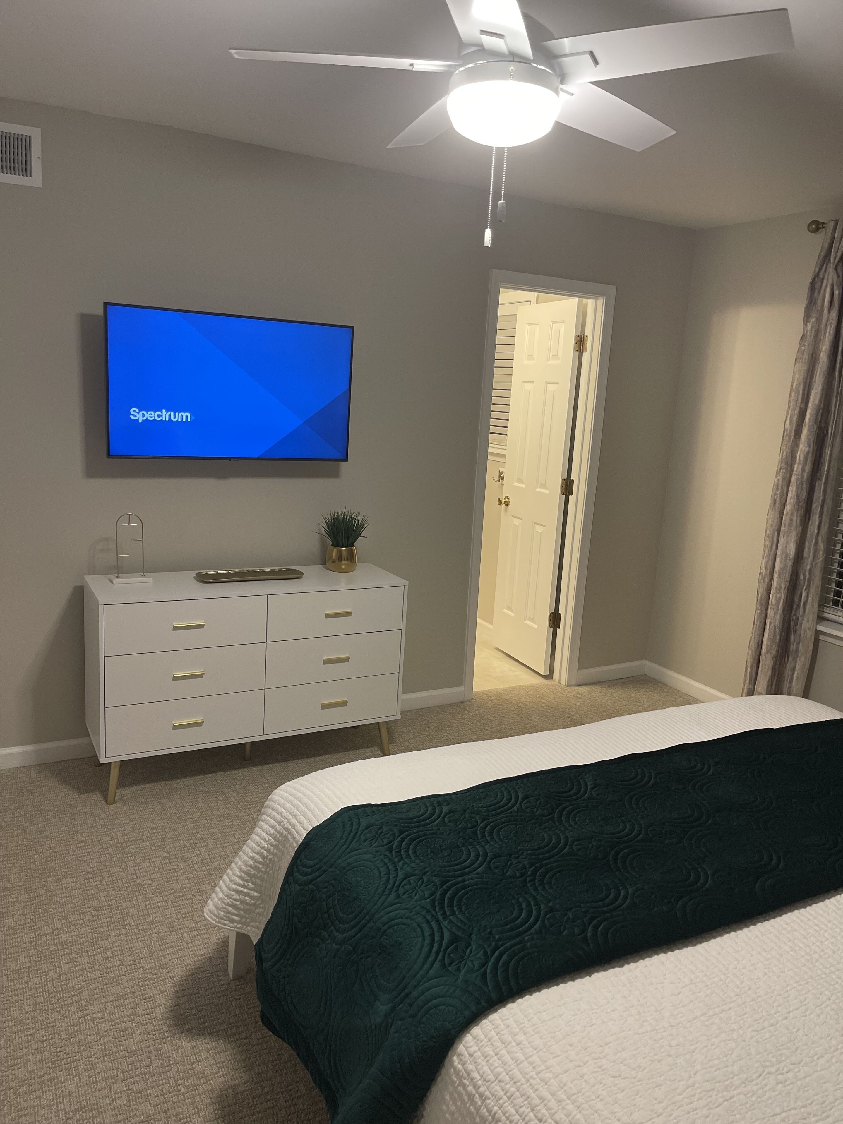 Une chambre à coucher comprenant un lit, une commode, un téléviseur diffusant l’émission « Spectrum », un ventilateur de plafond et une porte donnant sur une autre pièce.