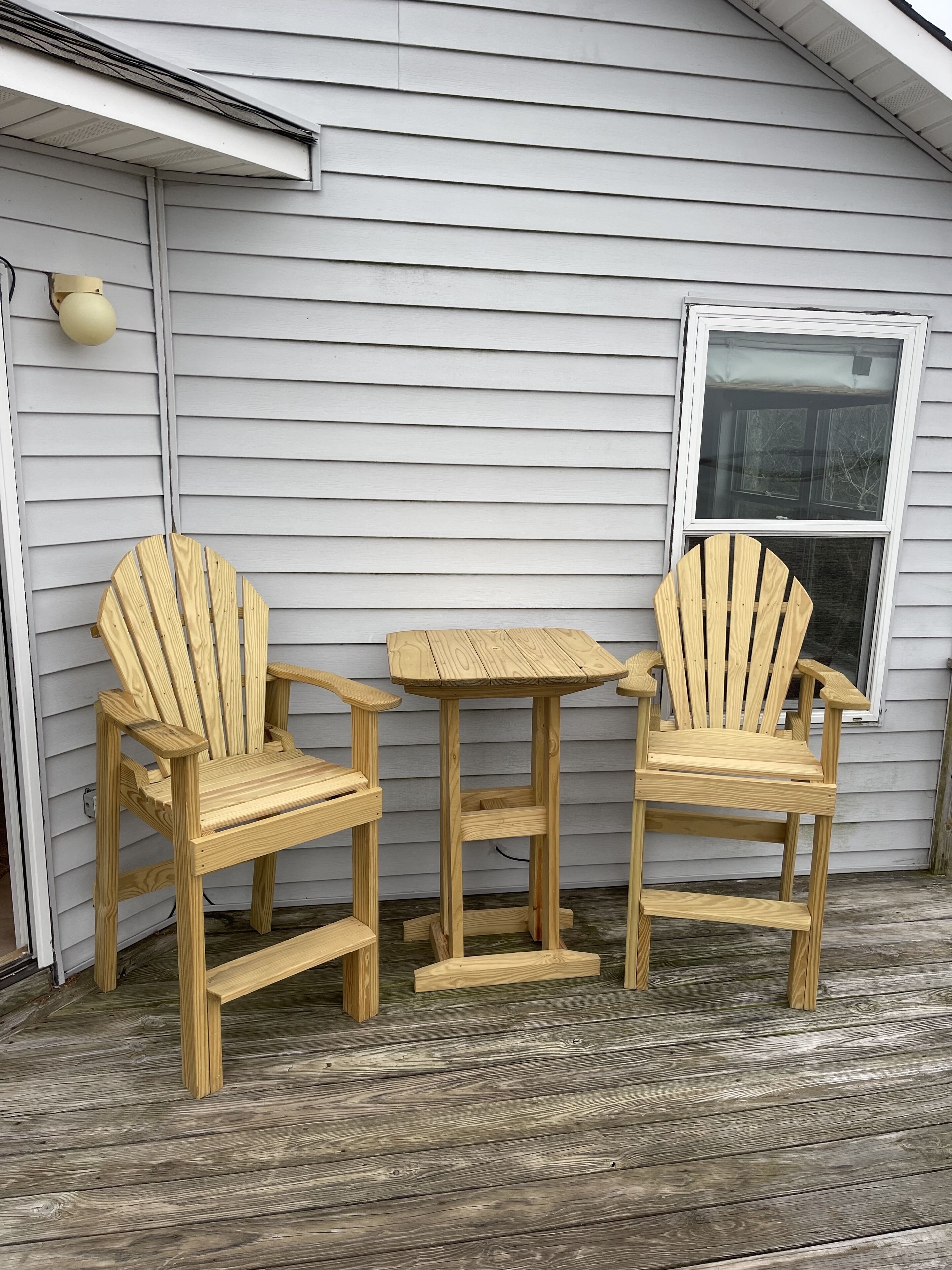 Two wooden Adirondack chairs and a small square table on a wooden deck in front of a house with white siding and a window.