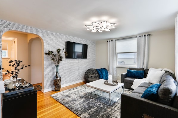 A modern living room with a white brick wall, a flat-screen TV mounted above a sofa, and a wooden floor.