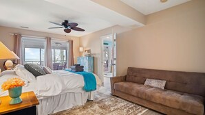 A hotel room with a bed, a brown sofa, a desk with a lamp, a window with curtains, and a ceiling fan.