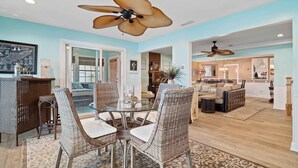 A spacious living room with a glass-top dining table surrounded by wicker chairs, a ceiling fan, and a view into the adjacent kitchen area.