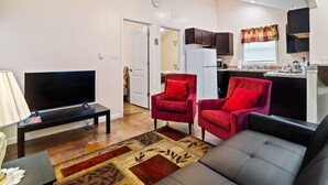 Ein Wohnzimmer mit Fernseher, zwei roten Sesseln, einer Couch, einem Deckenventilator und einer Küche im Hintergrund.