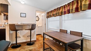 Ein Essbereich mit Holztisch und zwei Barhocker, ein Küchenbereich mit Granitarbeitsplatte sowie ein Fenster mit Jalousien und dekorativem Vorhang.
