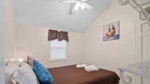 Ein Schlafzimmer mit Bett, Deckenventilator, Fenster mit Vorhängen, einem Gemälde und einem Heizkörper.