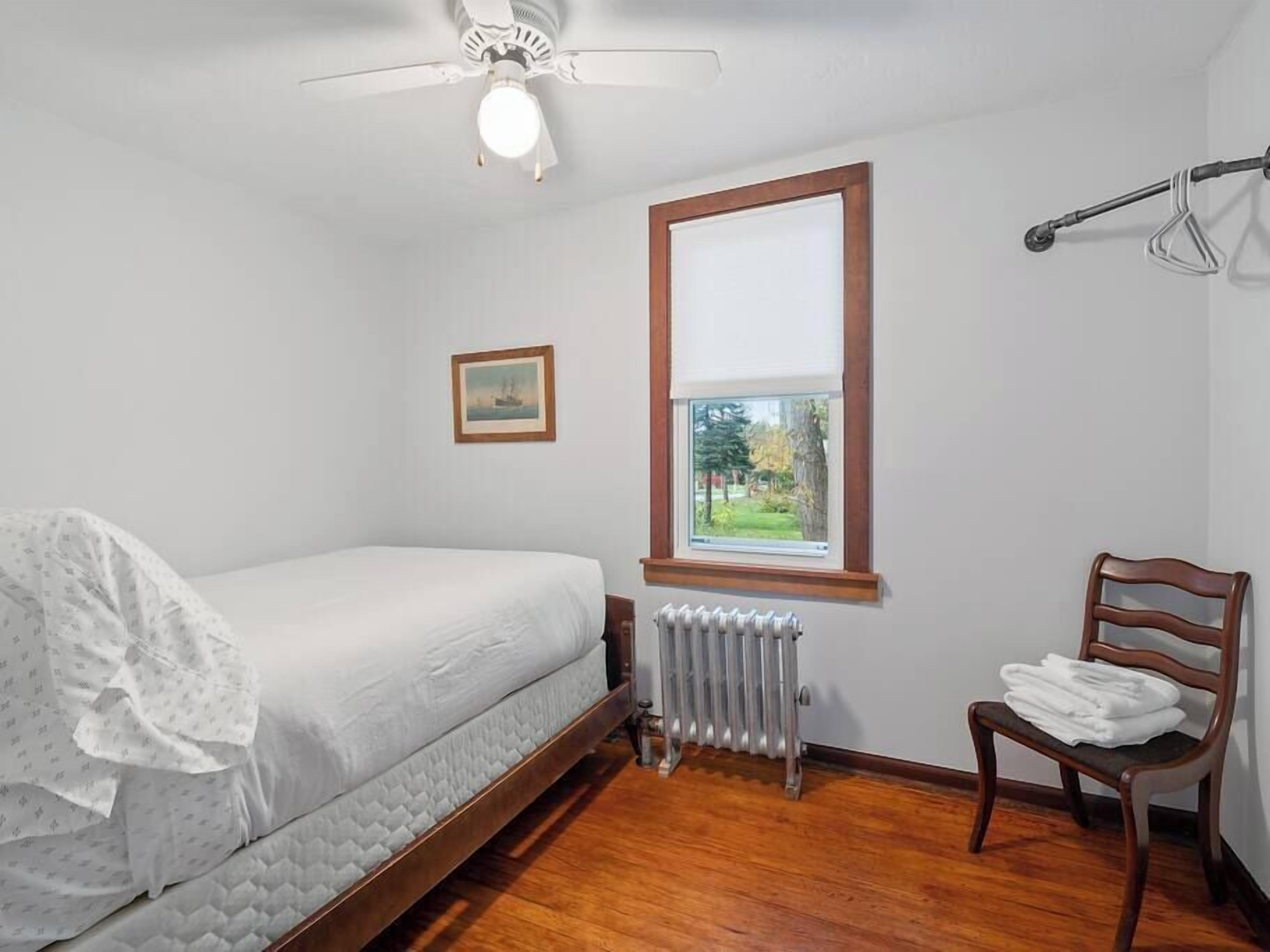 Une chambre à coucher avec un lit, un radiateur, une fenêtre donnant sur un paysage agréable, une chaise et un ventilateur de plafond.