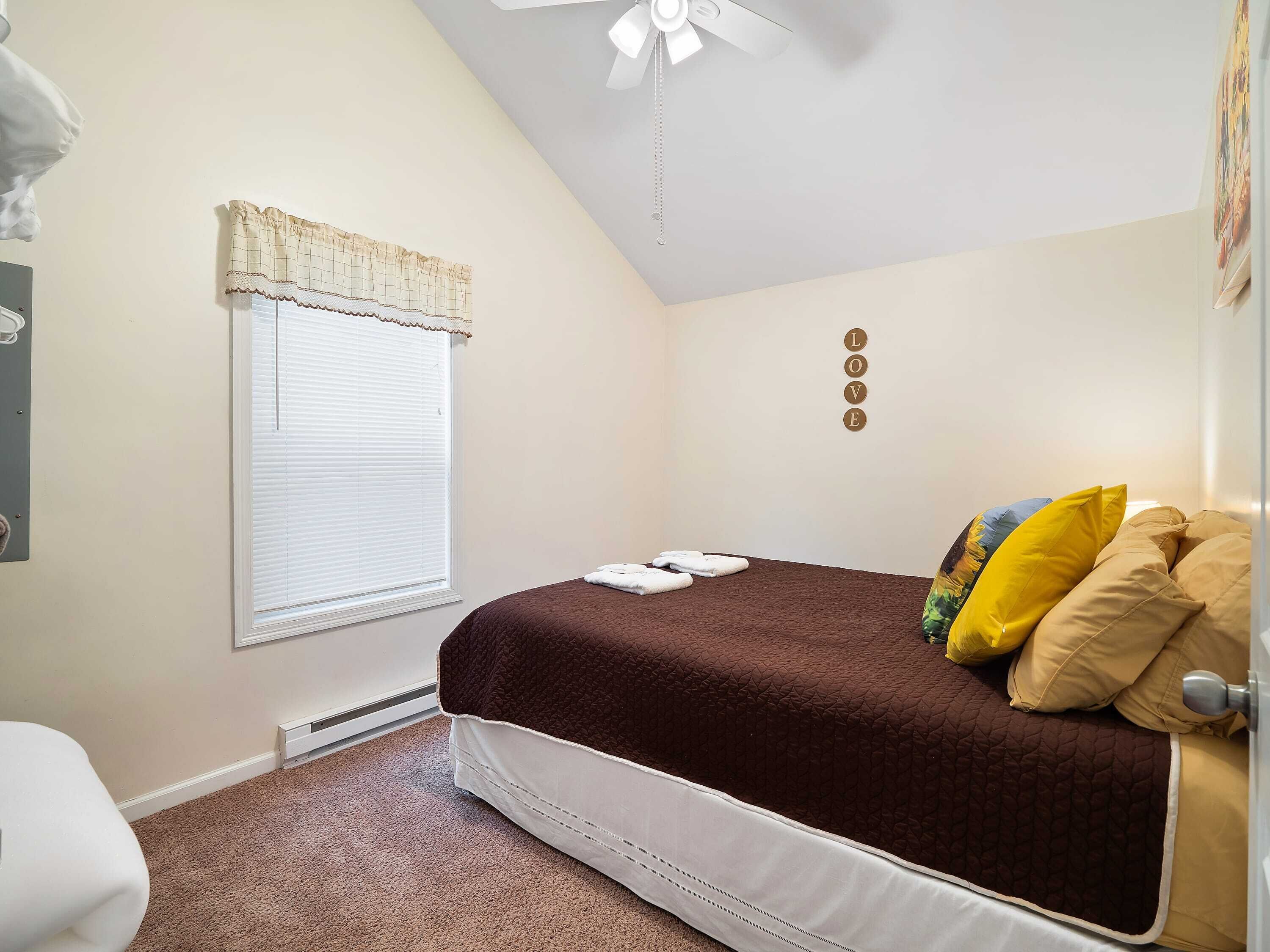 A bedroom with a bed, a ceiling fan, a window with blinds, and a wall-mounted artwork.