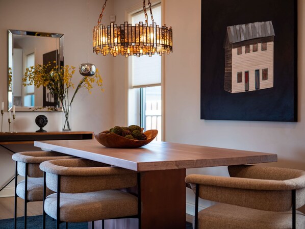 A modern dining area with a wooden table, cushioned benches, a decorative bowl, a hanging light fixture, a mirror, and a painting on the wall.