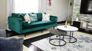 A living room with a green sofa, a round coffee table, and a flat-screen TV mounted on a stone-textured wall. - Topaz Villas (Lilongwe)