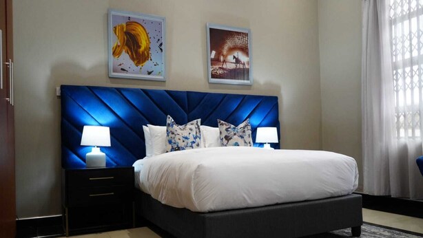A hotel room with a large bed, two bedside tables with lamps, a dark blue headboard, and two framed pictures on the wall. - Topaz Villas (Lilongwe)