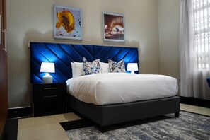 A hotel room with a large bed, two bedside tables with lamps, a dark blue headboard, and two framed pictures on the wall. - Topaz Villas (Lilongwe)