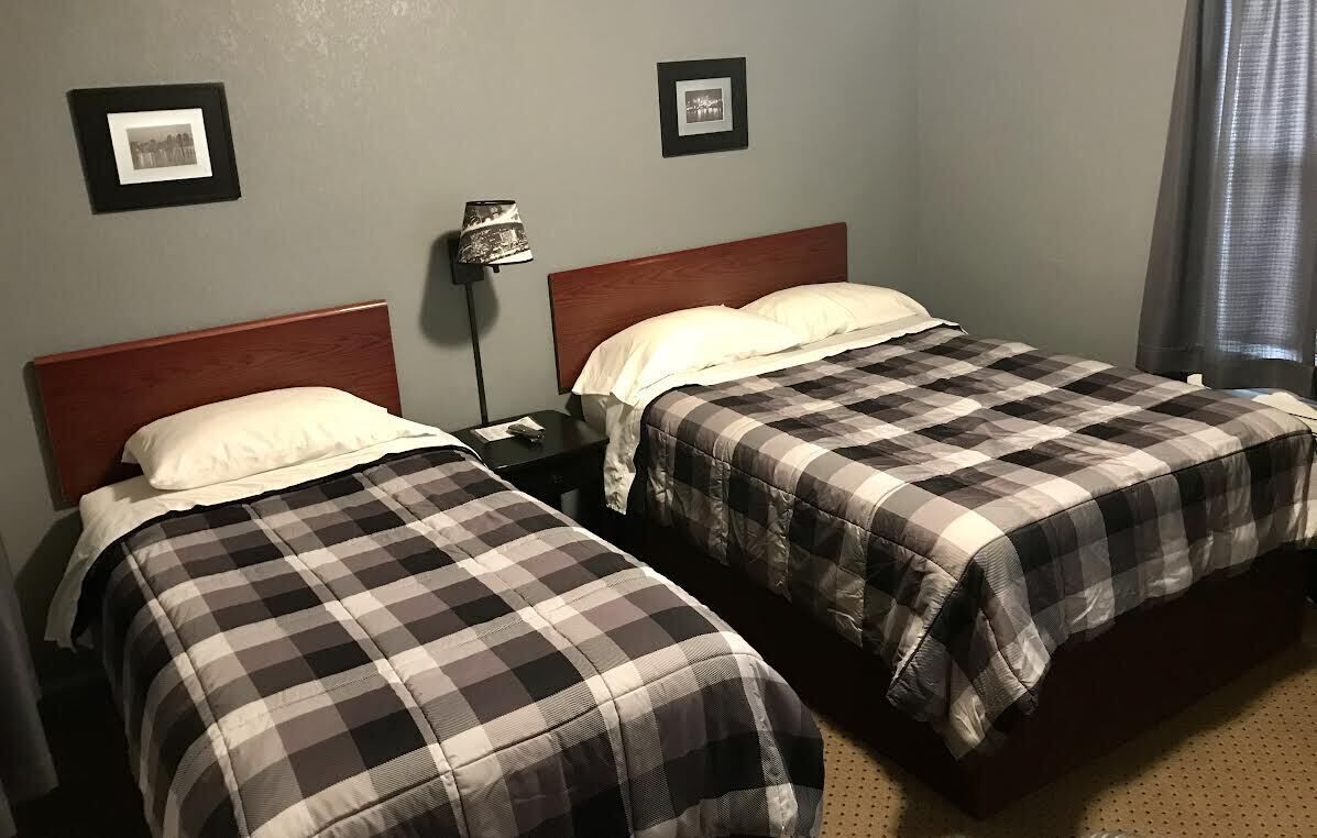 Ein Zimmer mit zwei Betten, beide mit karierten Bettdecken und weißen Kissen. An der Wand befinden sich gerahmte Bilder und eine Stehlampe.