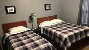 Ein Zimmer mit zwei Betten, beide mit karierten Bettdecken und weißen Kissen. An der Wand befinden sich gerahmte Bilder und eine Stehlampe.