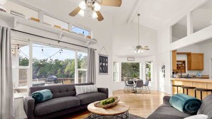 A spacious living area with a sofa, coffee table, and dining table. There are large windows and a view of greenery outside. - Canyon Cottage - 1 Acre w/Fire Pits & Game Room! (Canyon Lake)
