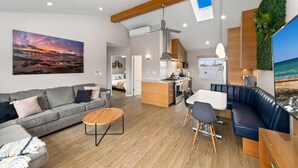 A modern living room with a sofa, coffee table, and dining area. There is a kitchen with a stove and microwave. A large window provides a view of the beach. - 7BD Family, Event Friendly 5min 2 Beach, Boardwalk (San Diego)