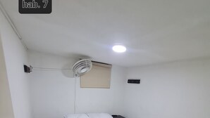 Una habitación con una cama, una mesita de noche, un reposapiés verde, un ventilador de techo y una ventana con persiana.