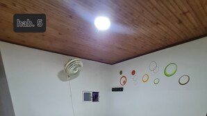 Una habitación con una cama, un banco verde, un ventilador, una ventana y adornos en la pared.