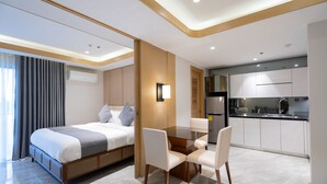 A modern hotel room with a bed, a dining table with chairs, a kitchen area, and a bathroom.