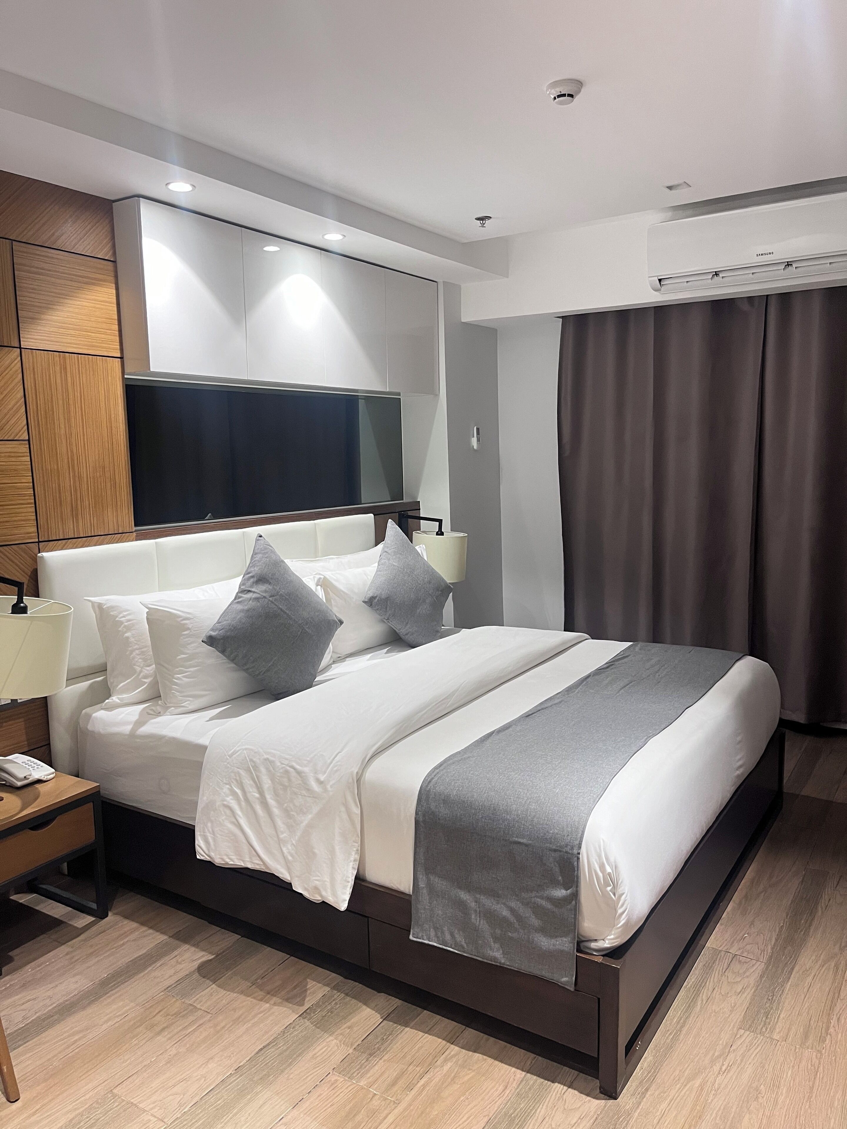 A modern hotel room with a large bed, a flat-screen TV, a bedside table with a lamp, and a built-in air conditioning unit.