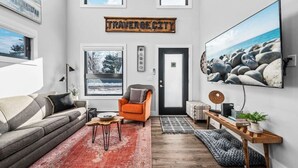 A modern living room with a grey sofa, an orange armchair, a wooden coffee table, and a large TV mounted on the wall.