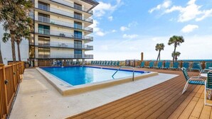 A pool area with a wooden deck, lounge chairs, and a building with balconies.