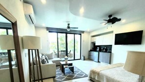 A modern living room with a sofa, coffee table, and a dining area. There is a kitchen with a TV and a ceiling fan. - Villa Dahlia, Luxury Studio, steps from the beach! (San Francisco)