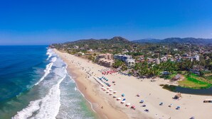 A coastal beach with a long stretch of sand, a row of beach umbrellas, and buildings in the background. - Villa Dahlia, Luxury Studio, steps from the beach! (San Francisco)