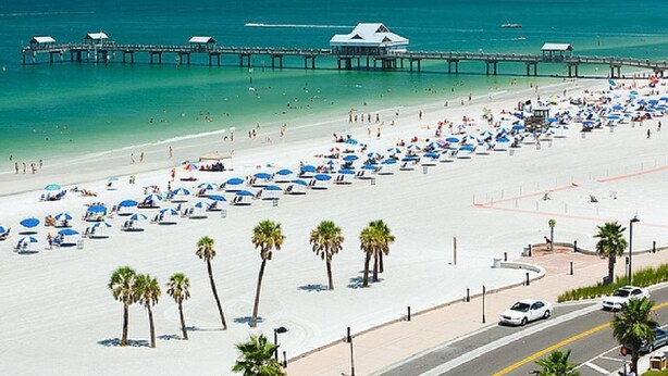 Een strand met een pier die zich uitstrekt over de oceaan, veel parasols en palmbomen.