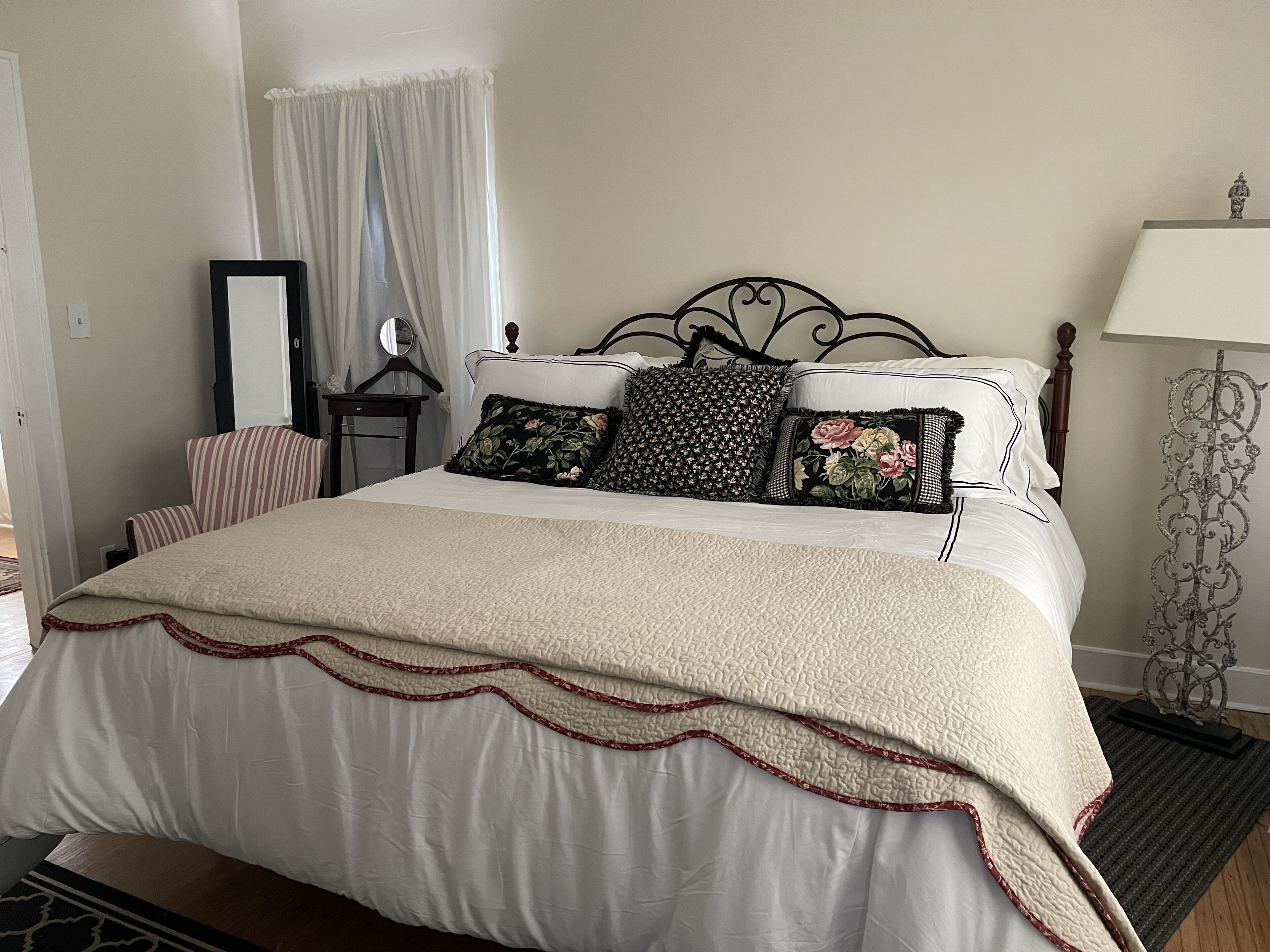 Una cama bien hecha con cubrecama acolchado y almohadas decorativas, una mesita de noche con espejo y una lámpara sobre un pedestal.