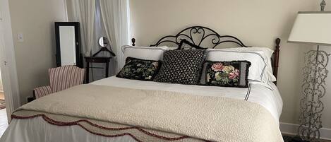 Una cama bien hecha con cubrecama acolchado y almohadas decorativas, una mesita de noche con espejo y una lámpara sobre un pedestal.