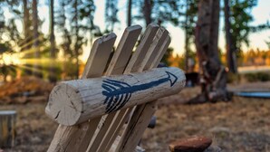 A rustic wooden chair with a directional arrow painted on it, set against a backdrop of trees and a wooden post.