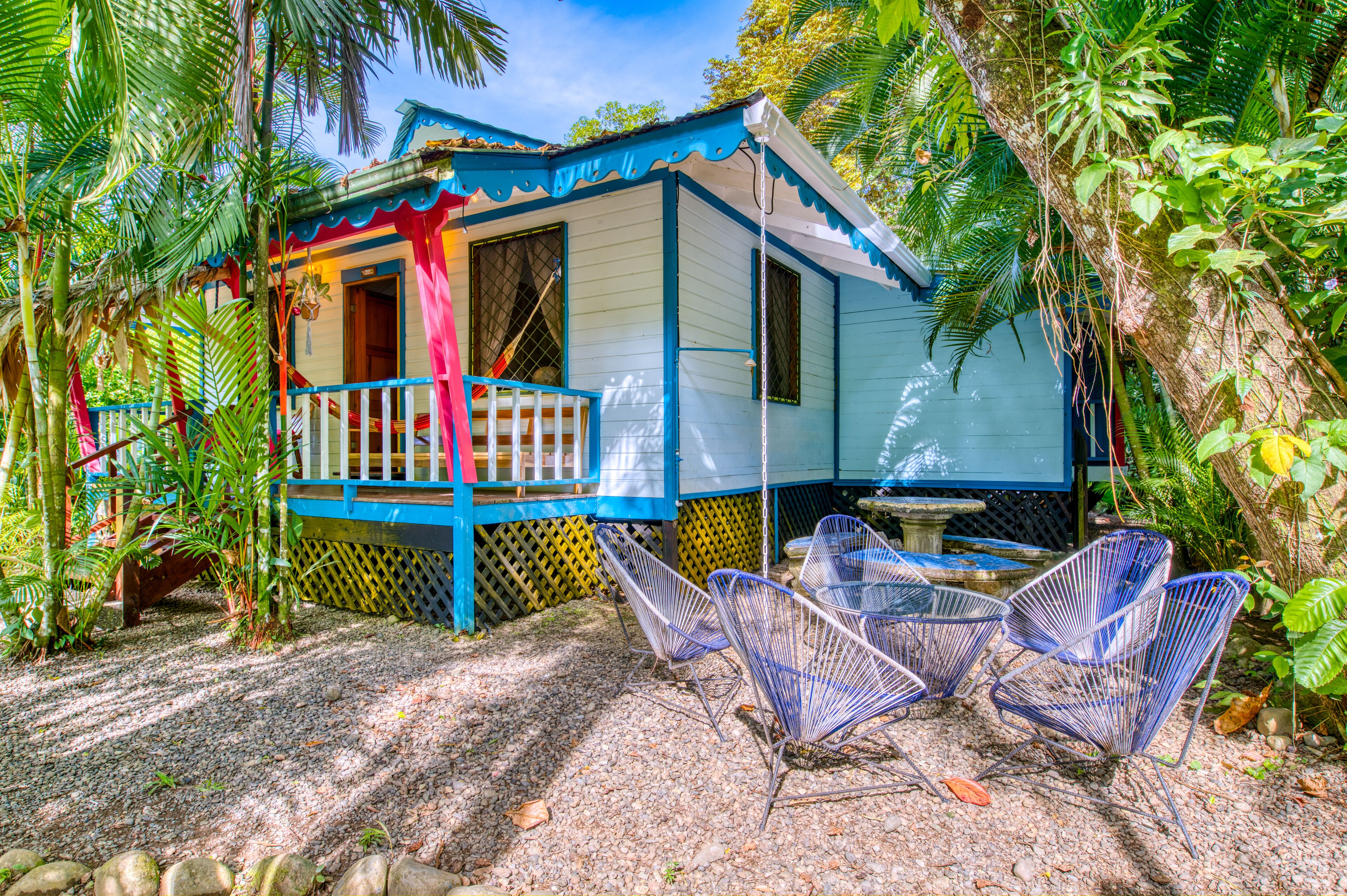 A small, colorful house with a porch and chairs, surrounded by lush greenery.