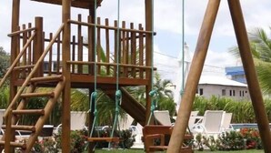 A wooden play structure with a ladder and slides, surrounded by greenery and a building in the background. - Miramar Laguna Beach (Ipojuca)