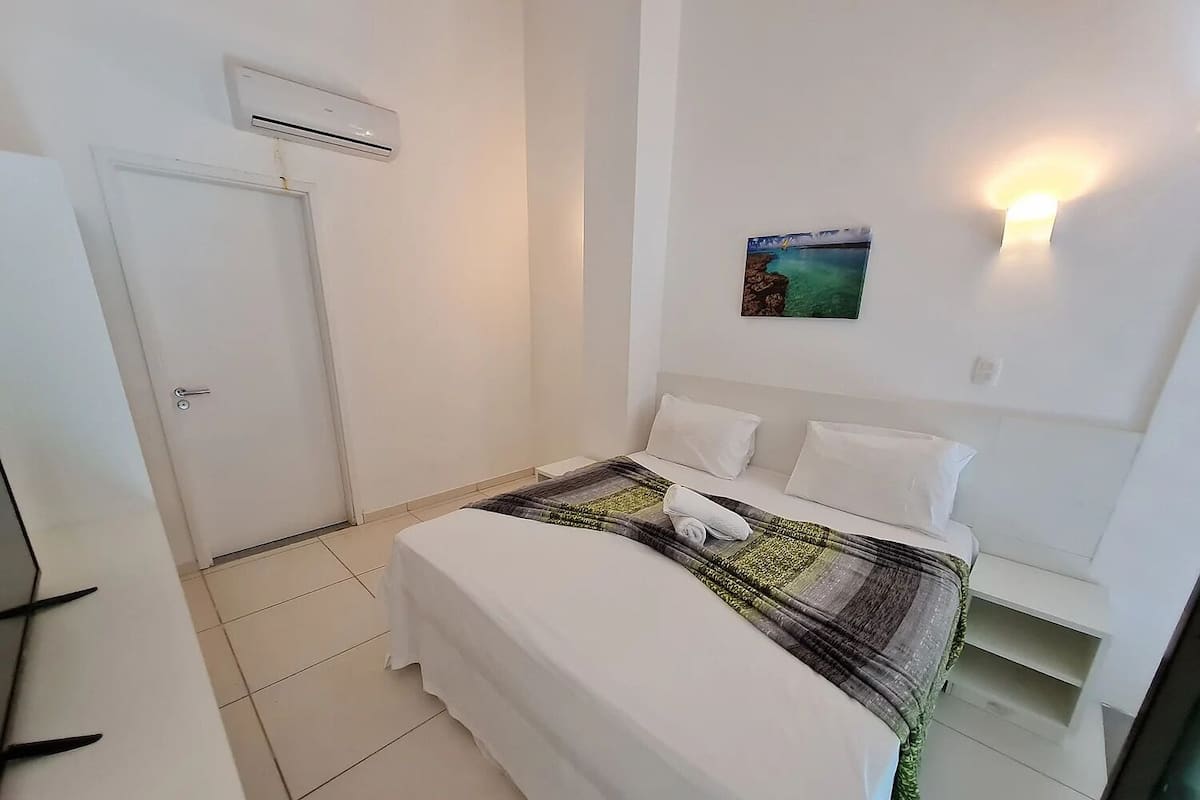 A hotel room with a bed, a wall-mounted air conditioner, a door, and a small shelf.