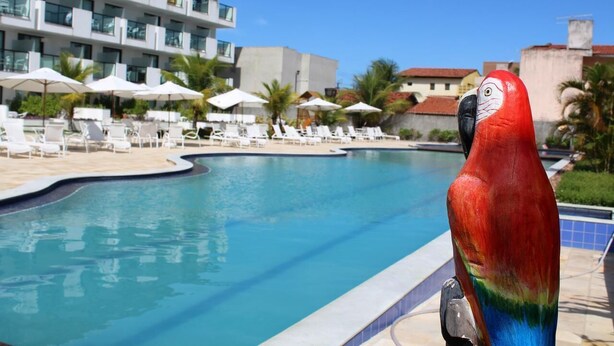 A pool area with lounge chairs and umbrellas, a modern building with balconies, and a parrot sculpture. - Miramar Laguna Beach (Ipojuca)