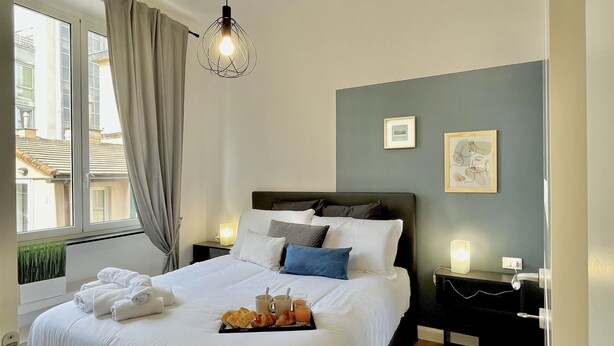 A modern bedroom with a large bed, bedside tables, a hanging light fixture, and a window with curtains. - Happy Family Apartments Lanterna (Genoa)