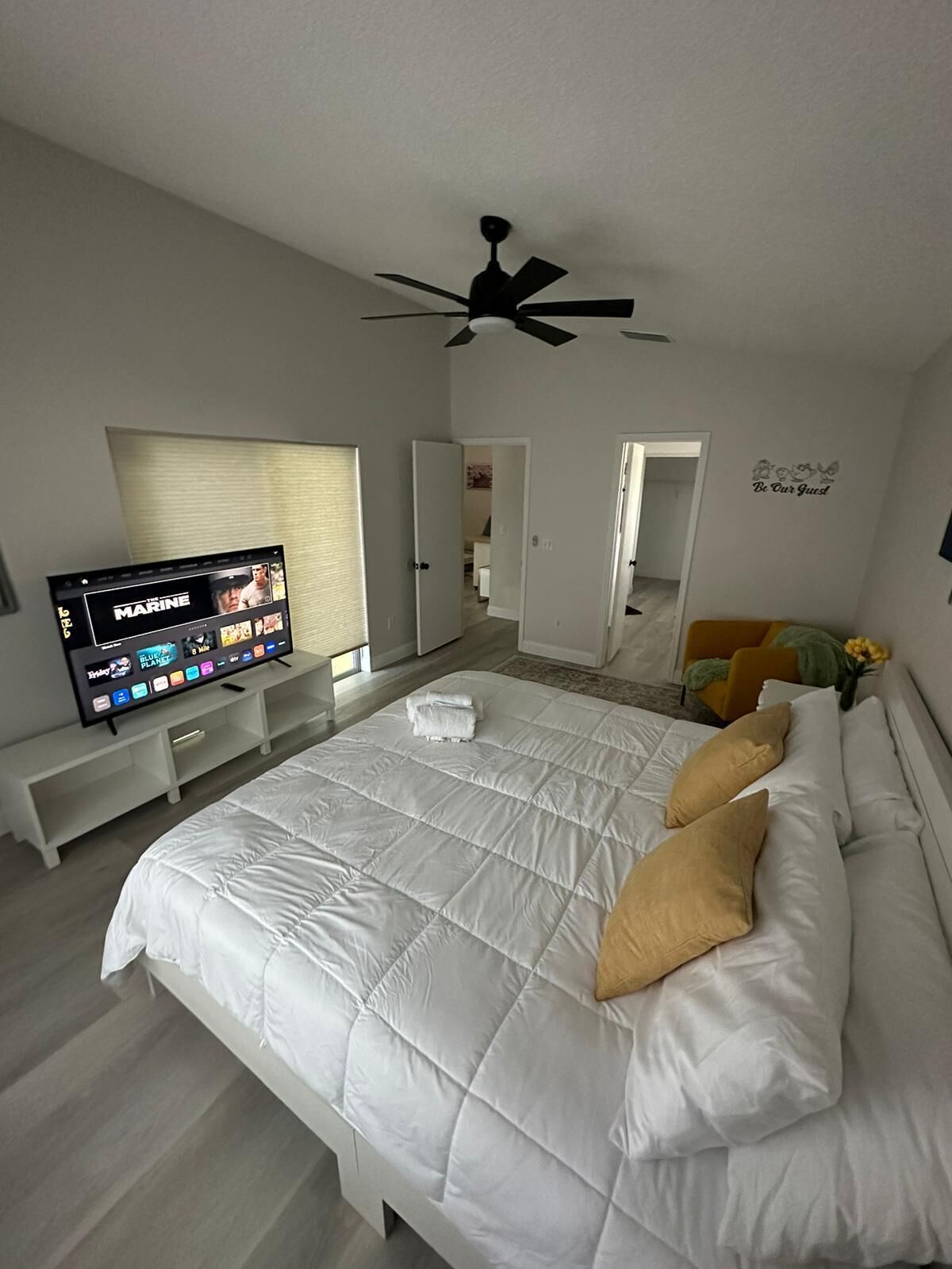 A modern bedroom with a large bed, a ceiling fan, a TV on a stand, and a 'Be Our Guest' wall decal.