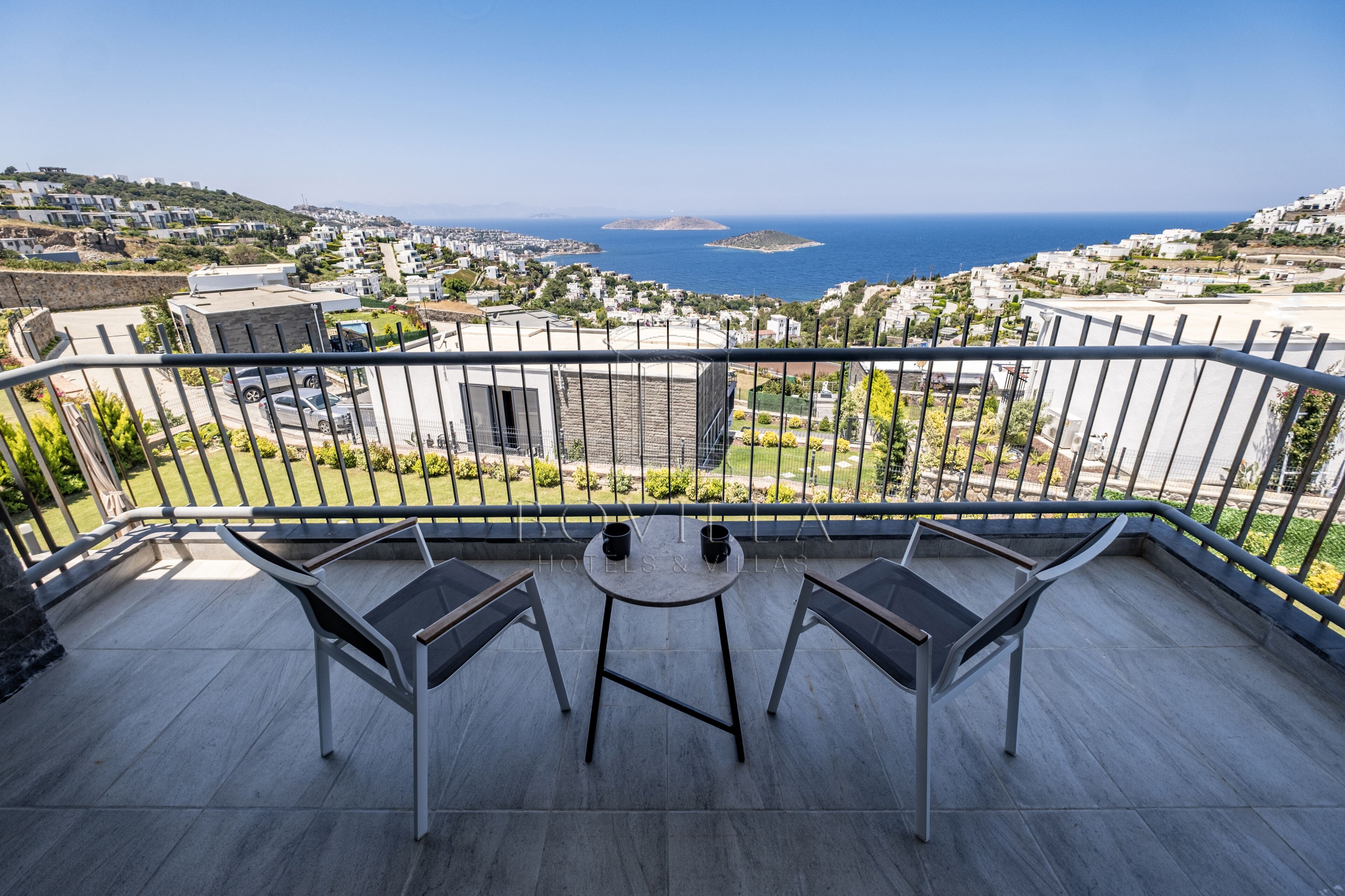 Un balcón con dos sillas y una mesita, con vistas a un pueblo costero con edificios y al mar.
