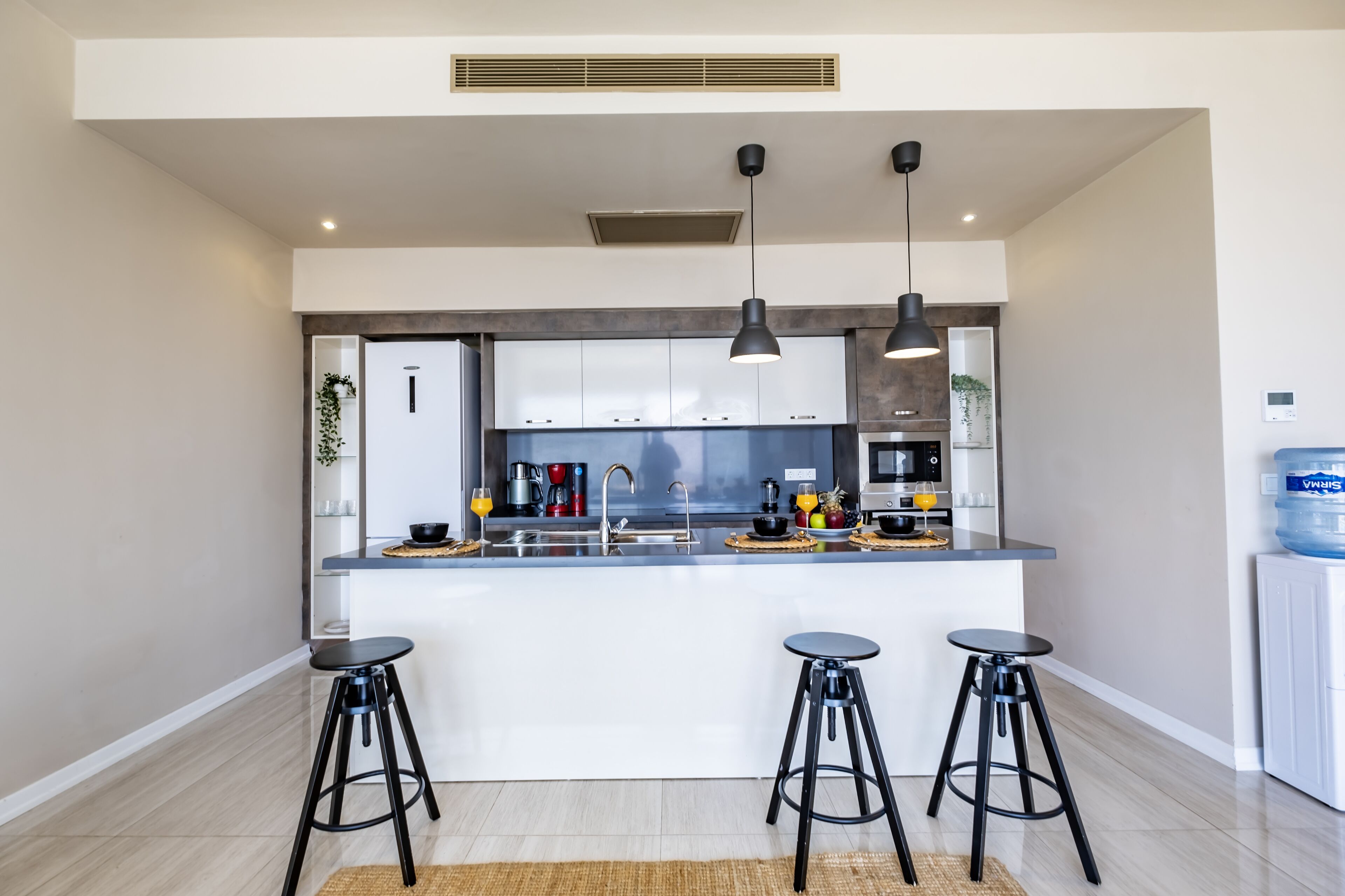 A modern kitchen with a central island, bar stools, pendant lighting, and a water dispenser.